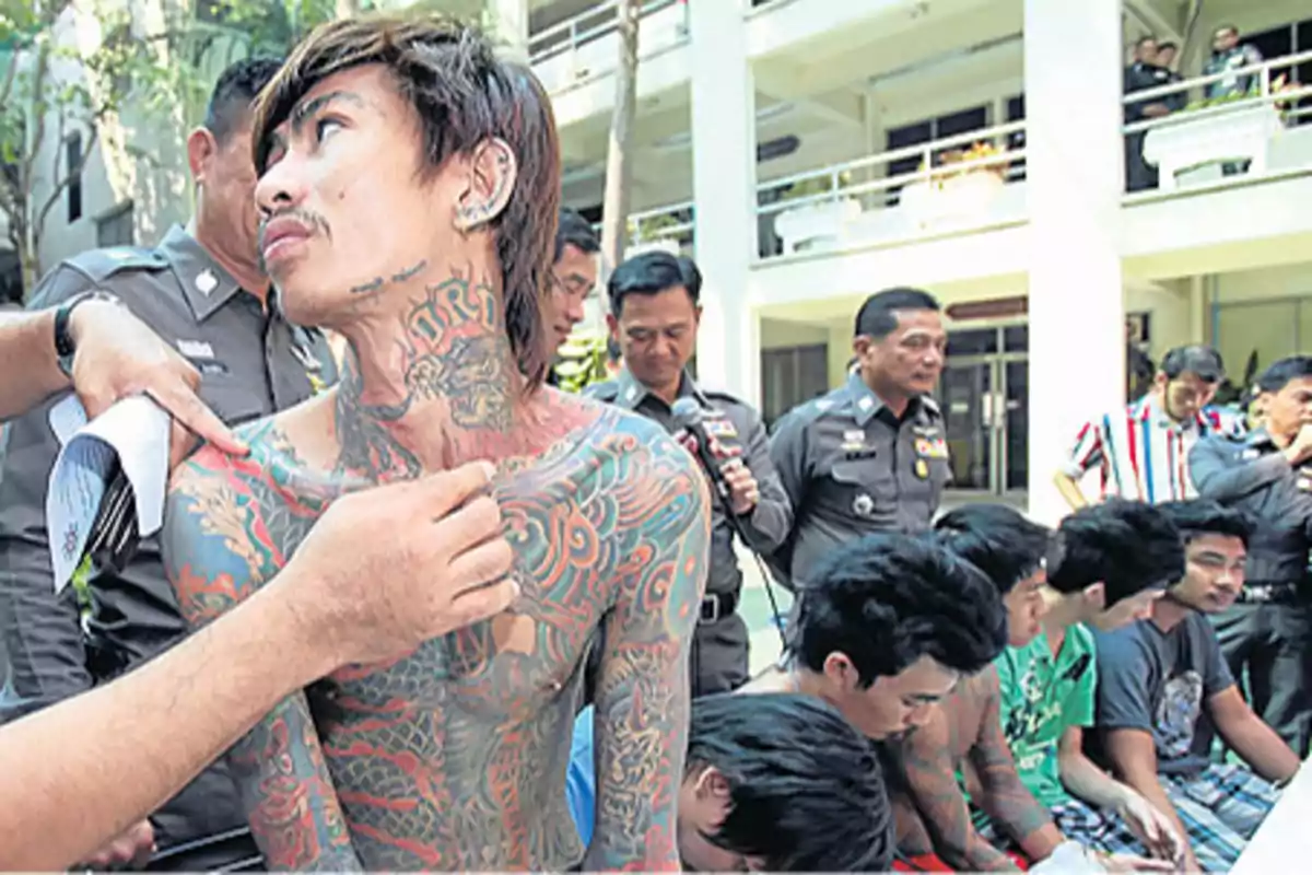 Un grupo de personas, algunas con tatuajes visibles, están sentadas mientras son observadas por varios oficiales de policía en un entorno al aire libre.