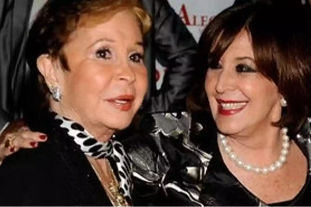 Dos mujeres sonrientes posan juntas en un evento formal, una lleva un collar de perlas y la otra un pañuelo con estampado de lunares. Dos mujeres sonrientes posan juntas en un evento formal, una lleva un collar de perlas y la otra un pañuelo con estampado de lunares.