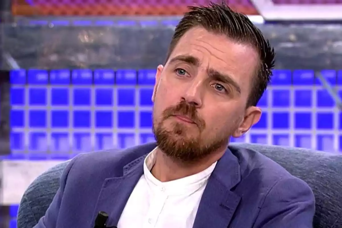 Un hombre con barba y cabello corto, vestido con una chaqueta azul y camisa blanca, está sentado en un estudio de televisión con un fondo de luces azules. Un hombre con barba y cabello corto, vestido con una chaqueta azul y camisa blanca, está sentado en un estudio de televisión con un fondo de luces azules.