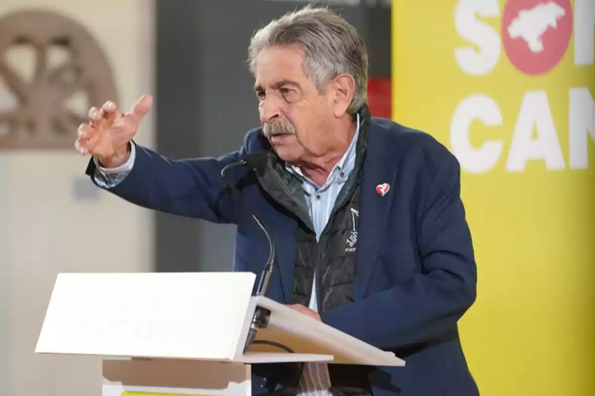 Un hombre mayor con bigote y cabello canoso está hablando en un podio con un micrófono, gesticulando con la mano, y detrás de él hay un cartel amarillo con texto parcialmente visible. Un hombre mayor con bigote y cabello canoso está hablando en un podio con un micrófono, gesticulando con la mano, y detrás de él hay un cartel amarillo con texto parcialmente visible.