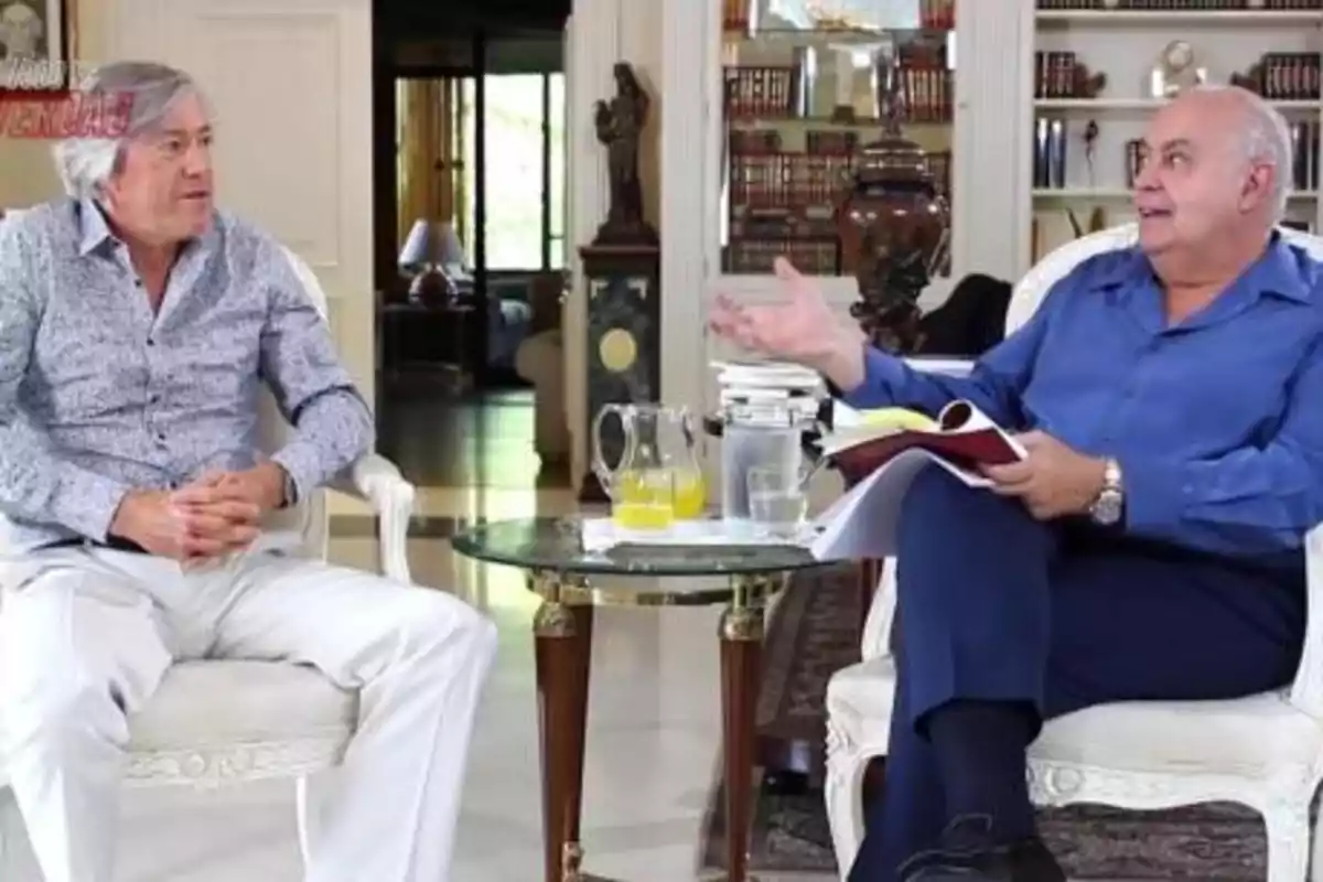 Dos hombres sentados en una sala, uno de ellos con una camisa azul y el otro con una camisa blanca, conversan mientras están frente a una mesa con una jarra de jugo y vasos.