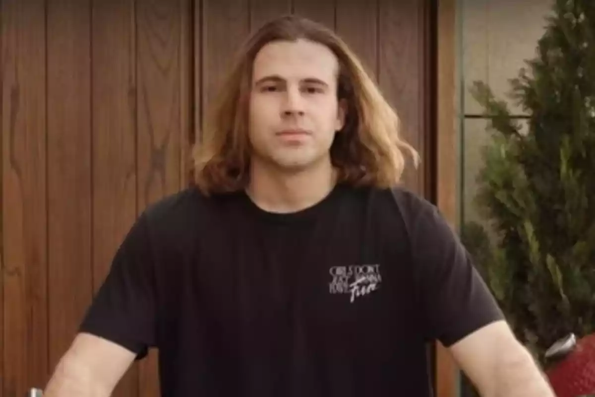 Un hombre con cabello largo y una camiseta negra está de pie frente a una puerta de madera.