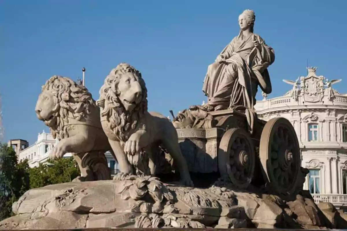 Estatua de una figura femenina en un carro tirado por dos leones en una plaza con edificios al fondo.