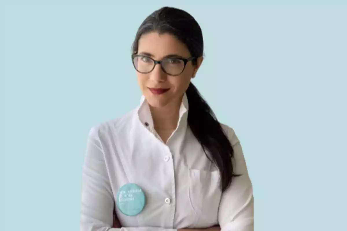 Mujer con bata blanca y gafas, con el cabello recogido y una insignia en el pecho, sobre un fondo azul claro.