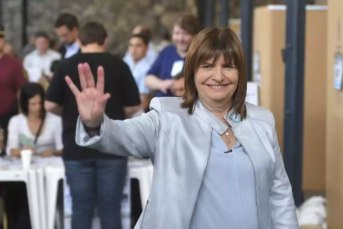 Una mujer sonriente con chaqueta clara levanta la mano en un lugar concurrido.