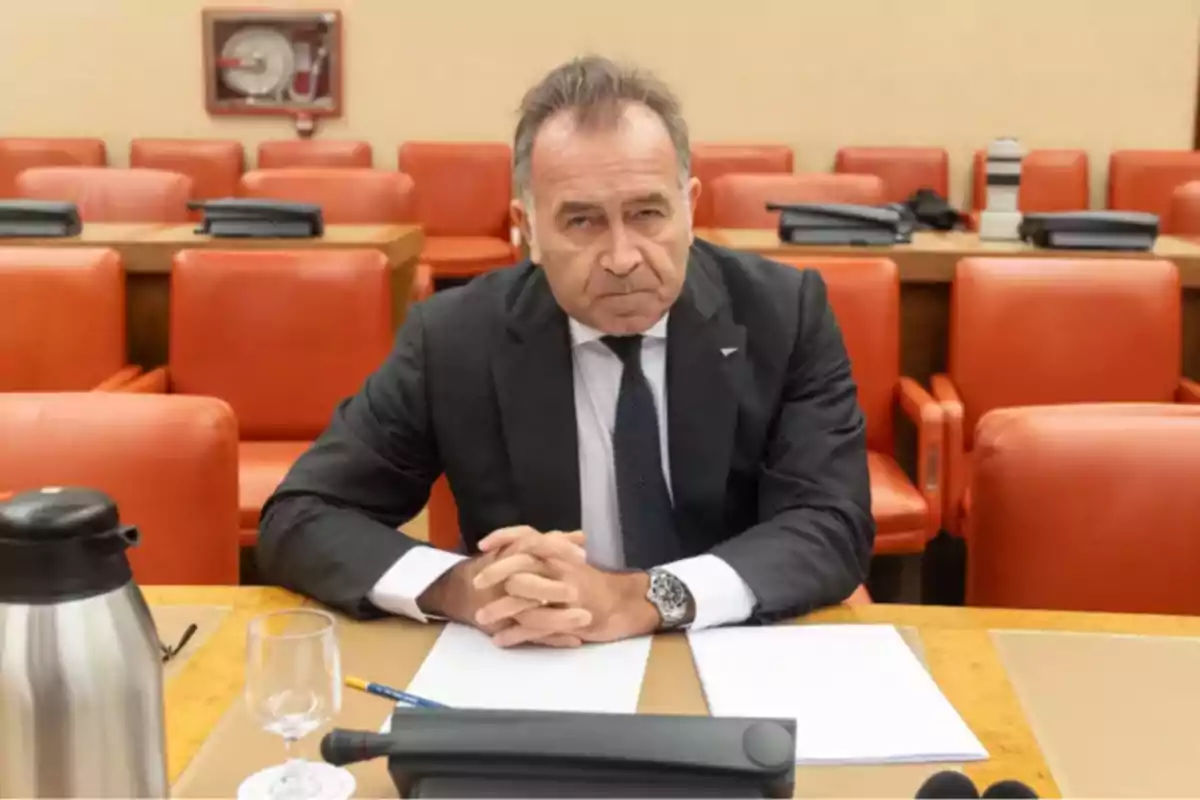 Un hombre de traje oscuro está sentado en una sala de conferencias con sillas rojas, frente a una mesa con documentos, un vaso y una jarra. Un hombre de traje oscuro está sentado en una sala de conferencias con sillas rojas, frente a una mesa con documentos, un vaso y una jarra.