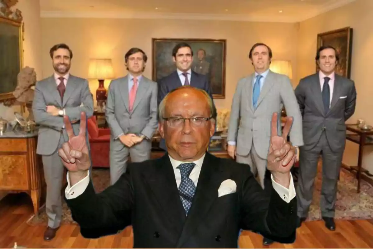 Un hombre mayor en primer plano con gafas y traje oscuro levanta ambas manos haciendo el signo de la victoria mientras cinco hombres jóvenes con trajes posan al fondo en una sala elegante