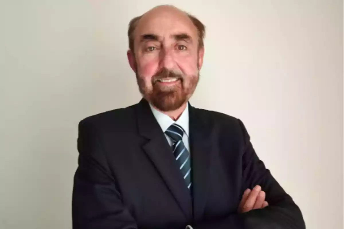 Un hombre con barba y traje oscuro sonríe con los brazos cruzados frente a un fondo claro. Un hombre con barba y traje oscuro sonríe con los brazos cruzados frente a un fondo claro.