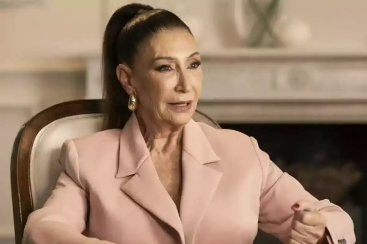 Mujer de cabello oscuro recogido con coleta alta, vestida con saco rosa claro, sentada en una silla elegante y hablando en un ambiente interior sofisticado Mujer de cabello oscuro recogido con coleta alta, vestida con saco rosa claro, sentada en una silla elegante y hablando en un ambiente interior sofisticado