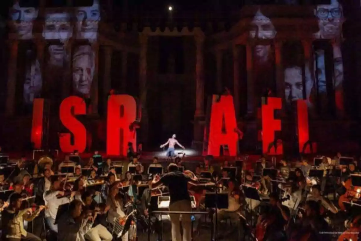Una orquesta sinfónica toca frente a grandes letras rojas que forman la palabra "ISRAEL", mientras rostros se proyectan en el fondo.