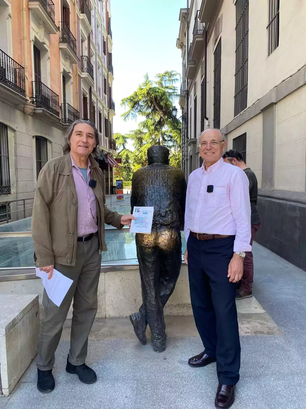 Dos hombres posan junto a una escultura en una calle rodeada de edificios, uno de ellos sostiene un papel.