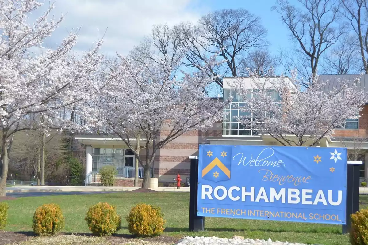 Un edificio escolar rodeado de árboles en flor con un cartel que da la bienvenida a Rochambeau, The French International School. Un edificio escolar rodeado de árboles en flor con un cartel que da la bienvenida a Rochambeau, The French International School.