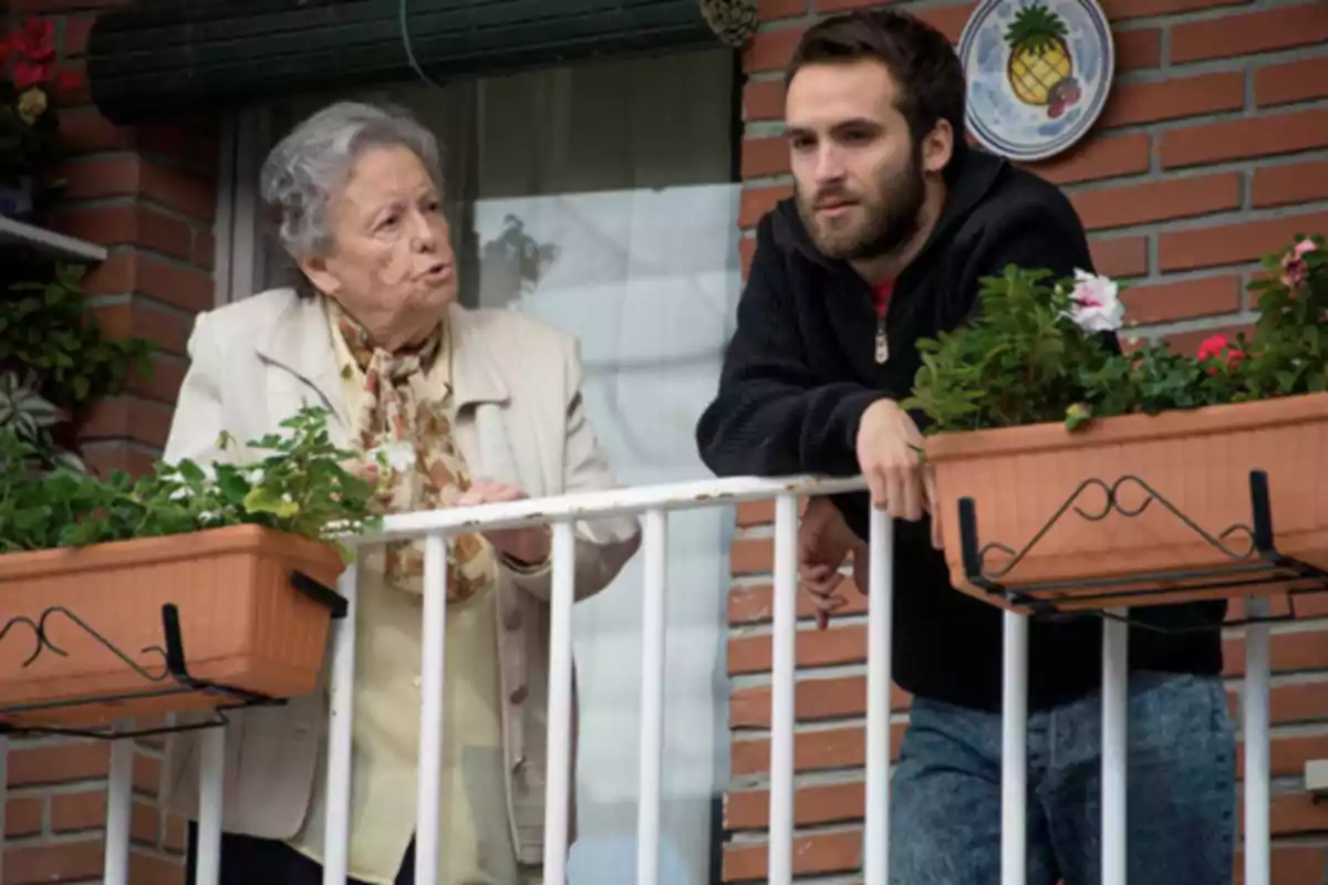 Una mujer mayor y un hombre joven conversan en un balcón adornado con macetas de flores.