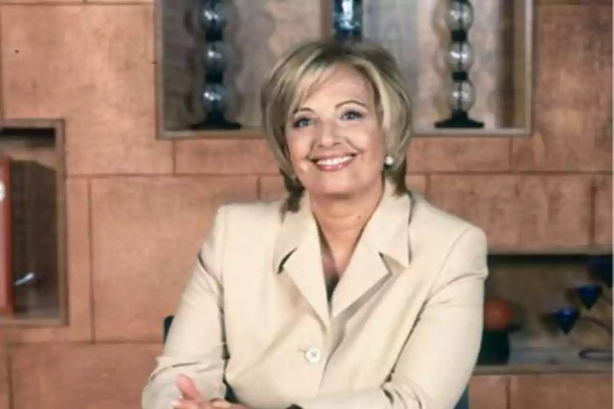 Una mujer sonriente con cabello rubio corto y vestida con un traje beige, sentada frente a una pared con estantes y decoraciones.