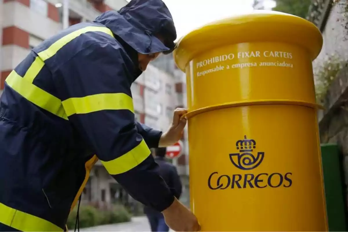 Persona con uniforme azul y amarillo depositando una carta en un buzón amarillo de Correos en la calle.