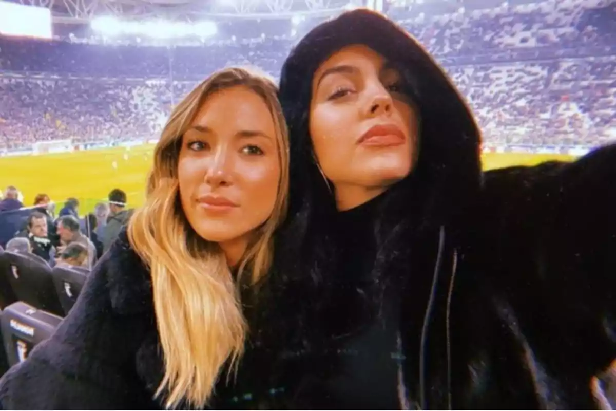 Dos mujeres posando para una selfie en un estadio de fútbol lleno de espectadores.