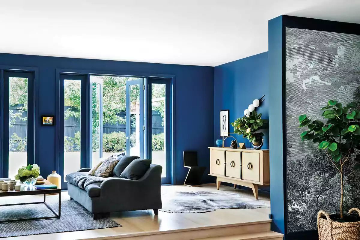 Sala de estar moderna con paredes azules, sofá gris, mesa de centro con decoración y puertas de vidrio que dan al jardín.