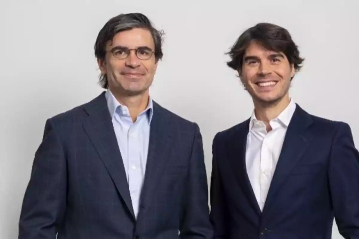 Dos hombres con trajes oscuros y camisas blancas posan sonriendo frente a un fondo claro.