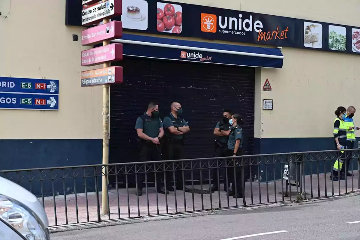 Un grupo de agentes de la Guardia Civil y personal de emergencias se encuentran frente a un supermercado Unide Market cerrado, junto a una señal de tráfico que indica direcciones hacia Madrid y Burgos.