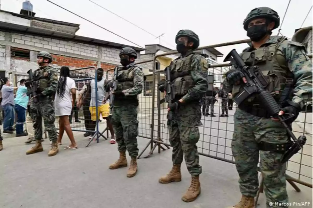 Soldados armados con uniformes de camuflaje y mascarillas negras están de pie en una fila frente a una reja, mientras varias personas se encuentran detrás de ellos en una zona urbana.