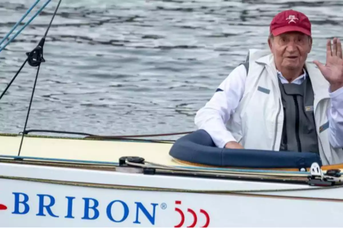 Un hombre mayor con gorra roja y chaleco blanco está sentado en un bote llamado "BRIBON" y saluda con la mano mientras navega en el agua.