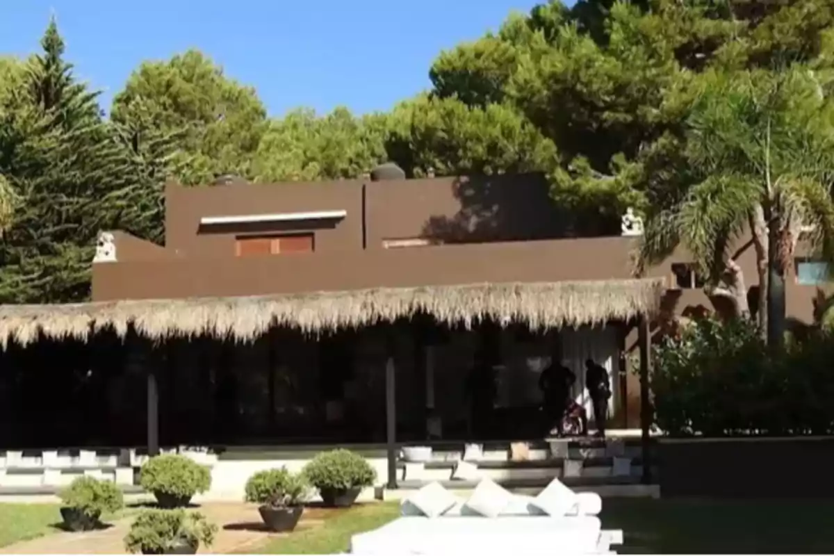 Una casa moderna de color marrón con un techo de paja, rodeada de árboles y vegetación, con un área de descanso al frente.