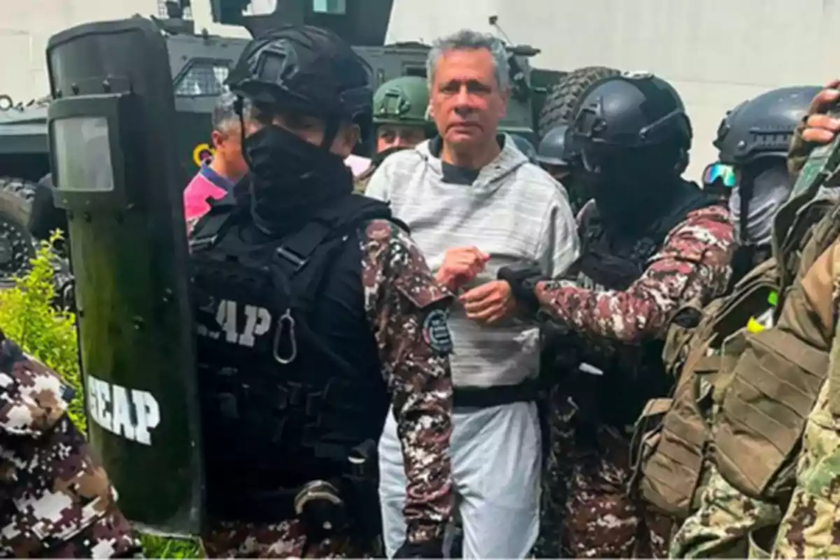 Un grupo de agentes de seguridad con equipo táctico detiene a un hombre de cabello canoso y suéter gris frente a un vehículo blindado.