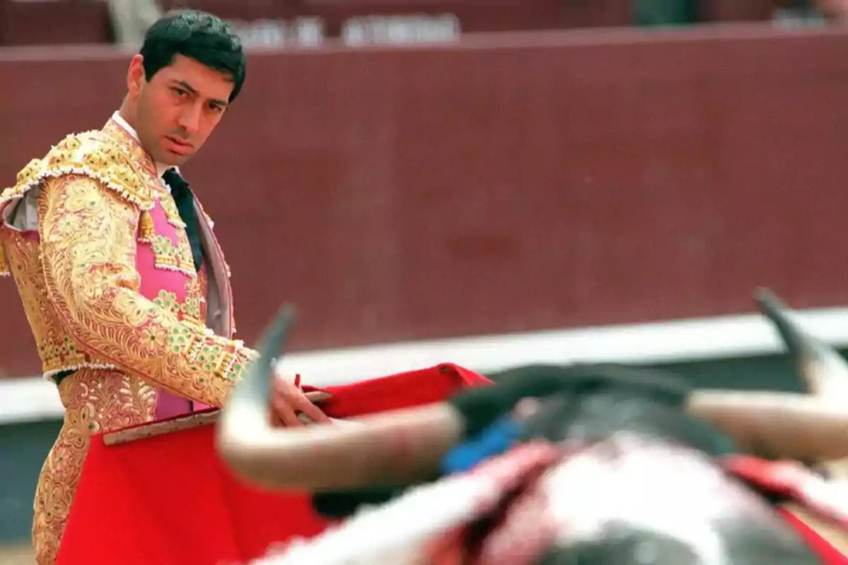 Un torero vestido con traje de luces dorado y rosa sostiene un capote rojo mientras observa al toro en una plaza de toros.