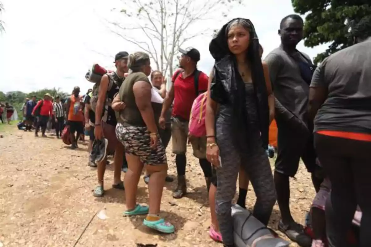 Un grupo de personas hace fila en un camino de tierra, algunas llevan mochilas y otras están vestidas con ropa casual.