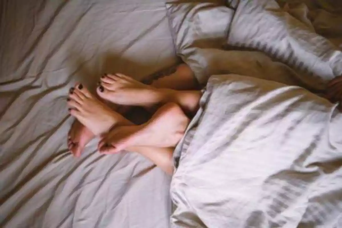 Pies de dos personas entrelazados bajo una sábana en una cama.