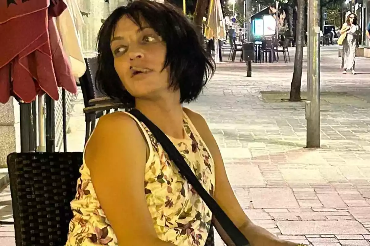 Una mujer con cabello corto y oscuro, vestida con una blusa de flores, está sentada en una silla en una calle iluminada por la noche.