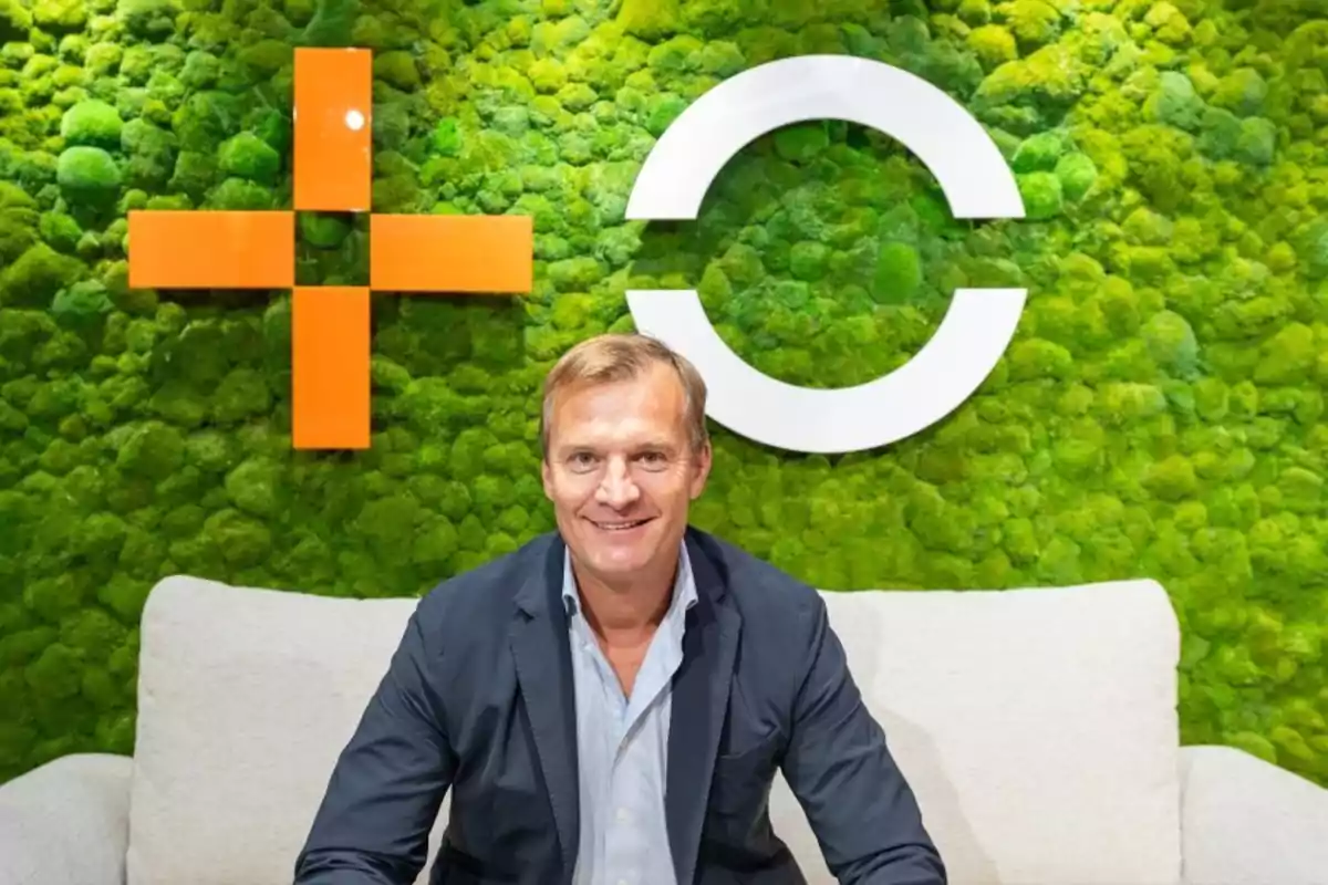 Hombre rubio con saco azul sentado en un sofá claro frente a una pared verde con símbolos geométricos naranja y blanco Hombre rubio con saco azul sentado en un sofá claro frente a una pared verde con símbolos geométricos naranja y blanco