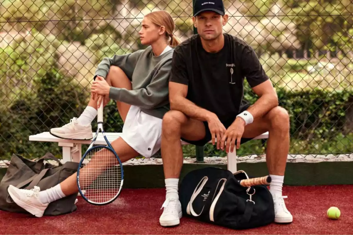 Dos personas sentadas en un banco de una cancha de tenis, una mujer con una raqueta y un hombre con una gorra, ambos con ropa deportiva y bolsas a su lado.