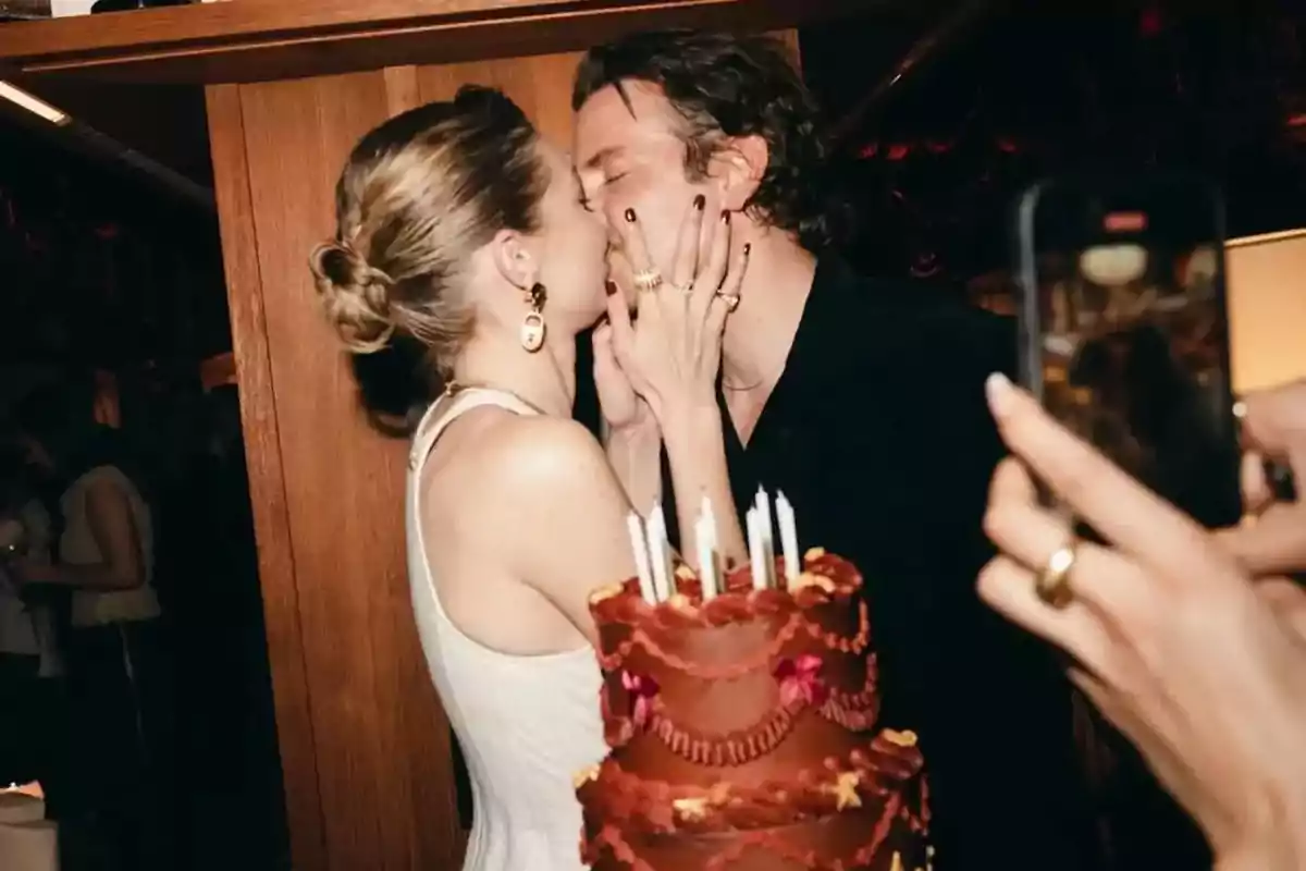 Una pareja se besa frente a un pastel de cumpleaños mientras alguien toma una foto con un teléfono móvil. Una pareja se besa frente a un pastel de cumpleaños mientras alguien toma una foto con un teléfono móvil.