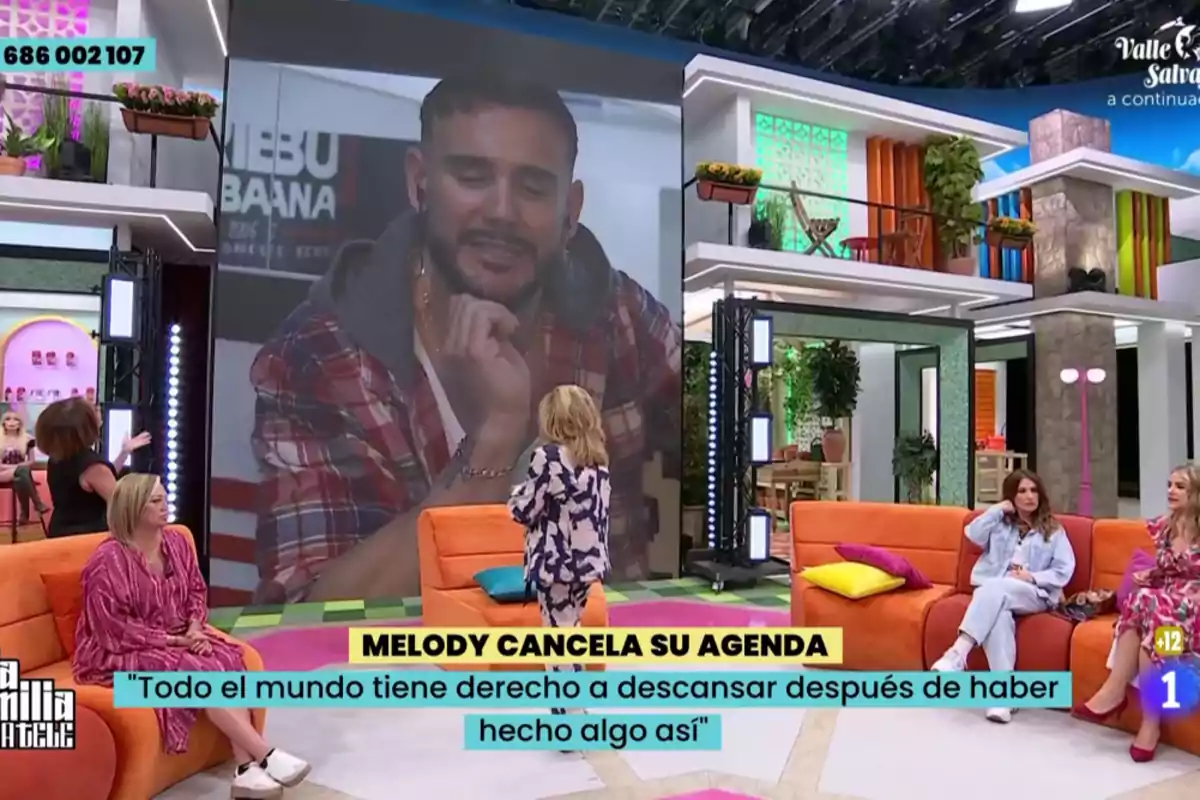 Un programa de televisión con varias personas sentadas en sofás naranjas y una gran pantalla mostrando a un hombre con camisa de cuadros, mientras en la parte inferior aparece un texto sobre la cancelación de la agenda de Melody Un programa de televisión con varias personas sentadas en sofás naranjas y una gran pantalla mostrando a un hombre con camisa de cuadros, mientras en la parte inferior aparece un texto sobre la cancelación de la agenda de Melody