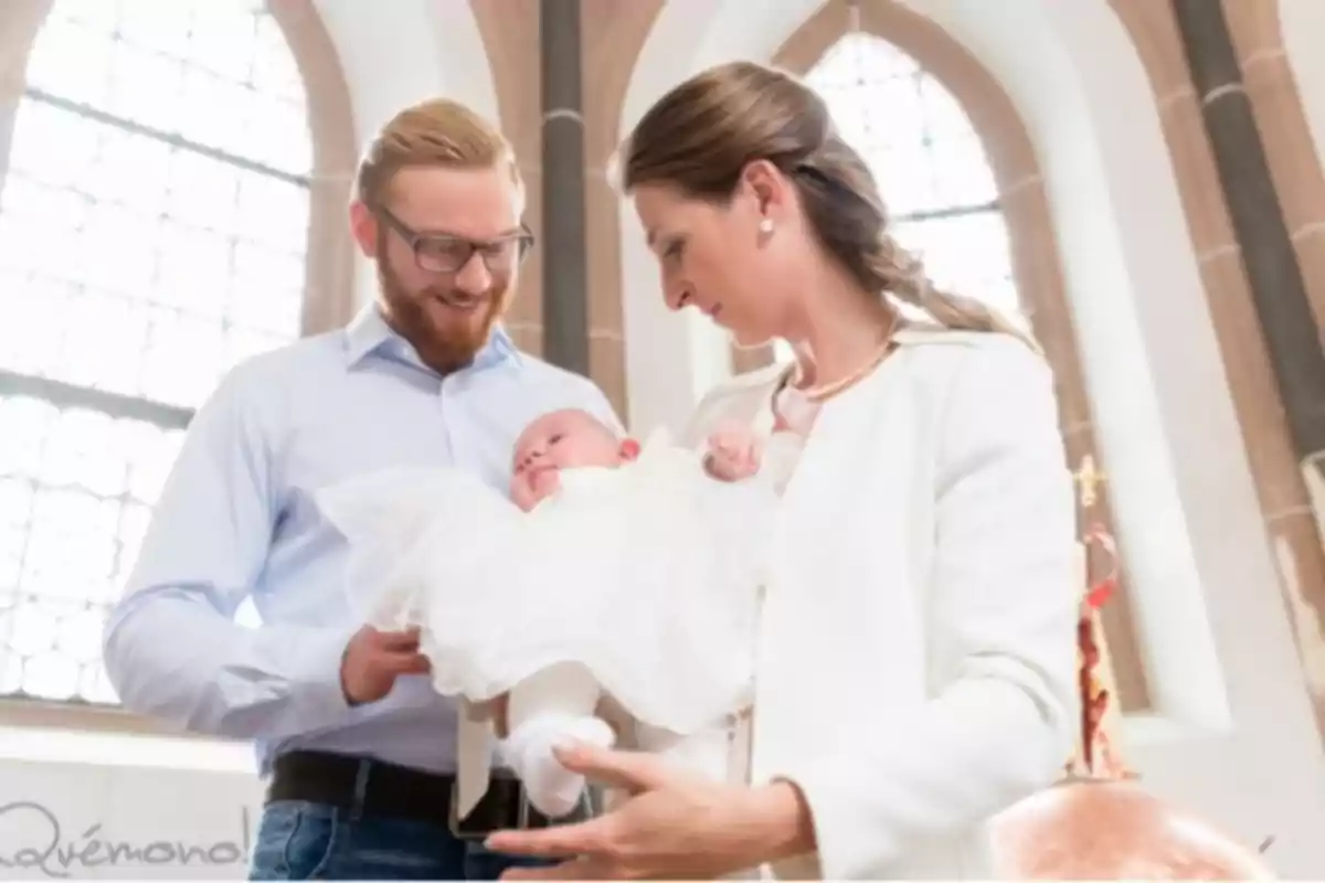 Una pareja sonriente sostiene a un bebé vestido de blanco en un entorno con grandes ventanas y columnas.