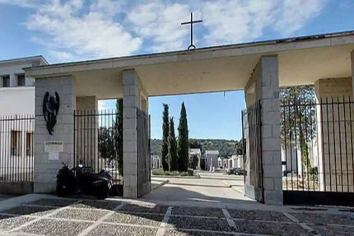 Entrada principal de un cementerio con una cruz en la parte superior y árboles en el fondo.