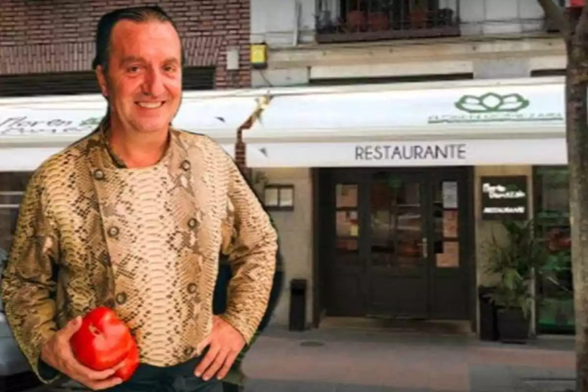 Un hombre sonriente con una chaqueta de estampado de serpiente sostiene un tomate frente a un restaurante.