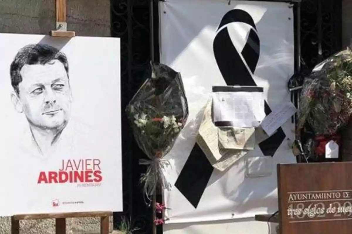 Un retrato de un hombre con el nombre "Javier Ardines" en un cartel, junto a un lazo negro y flores en un homenaje.