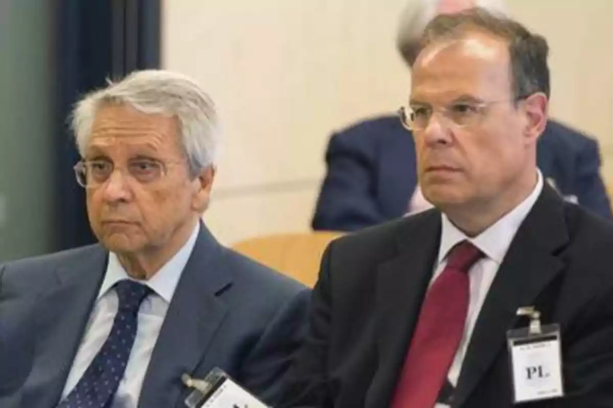 Dos hombres mayores con trajes formales y gafas, sentados en una conferencia o reunión, con expresiones serias y etiquetas de identificación en sus chaquetas.