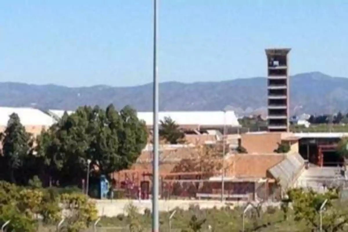 Vista de un edificio de ladrillo con una torre alta y árboles en primer plano, con montañas al fondo.