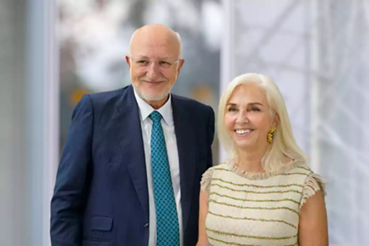 Una pareja sonriente posando juntos, el hombre lleva un traje azul y corbata verde, mientras que la mujer lleva un vestido claro con detalles dorados.