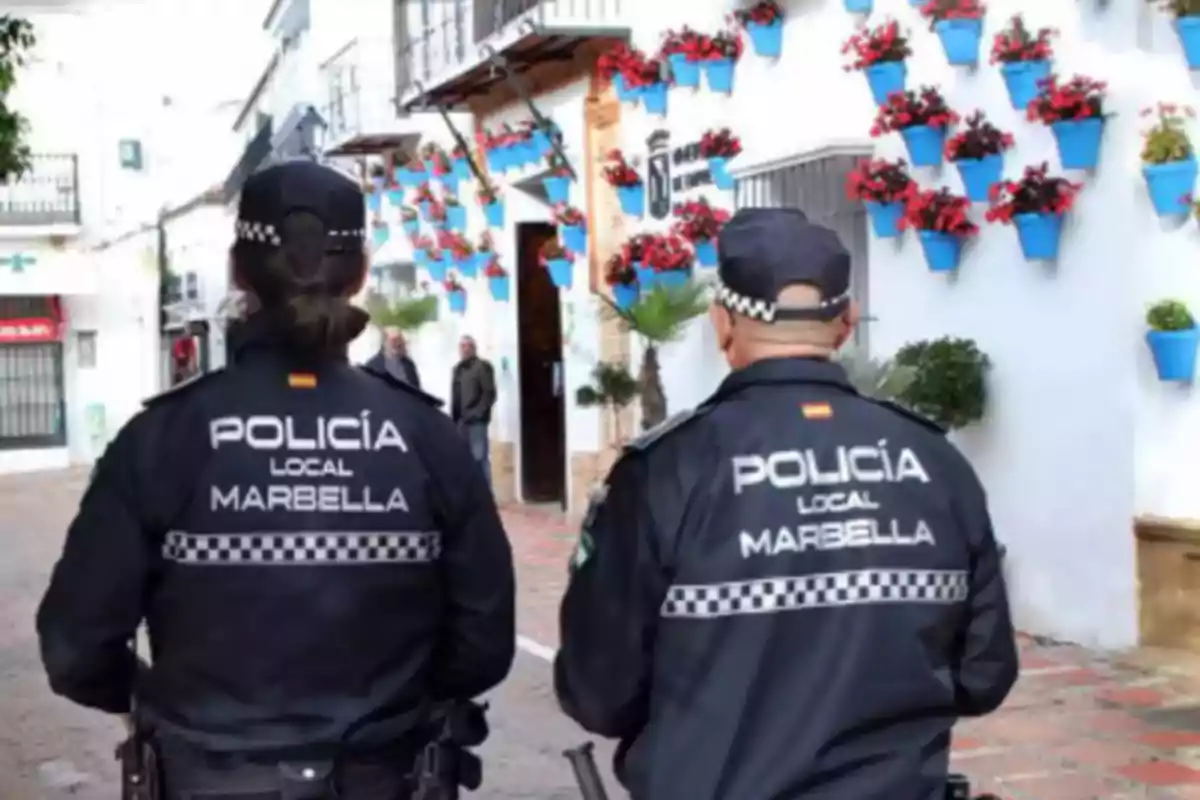 Dos agentes de la Policía Local de Marbella caminan por una calle adornada con macetas azules y flores rojas.