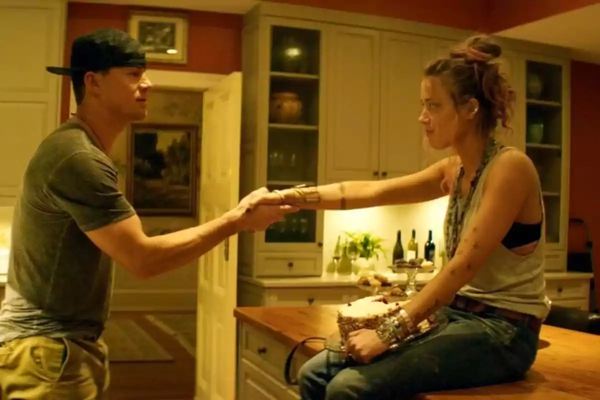 Una pareja se da la mano en una cocina mientras la mujer está sentada en la mesa y el hombre está de pie.