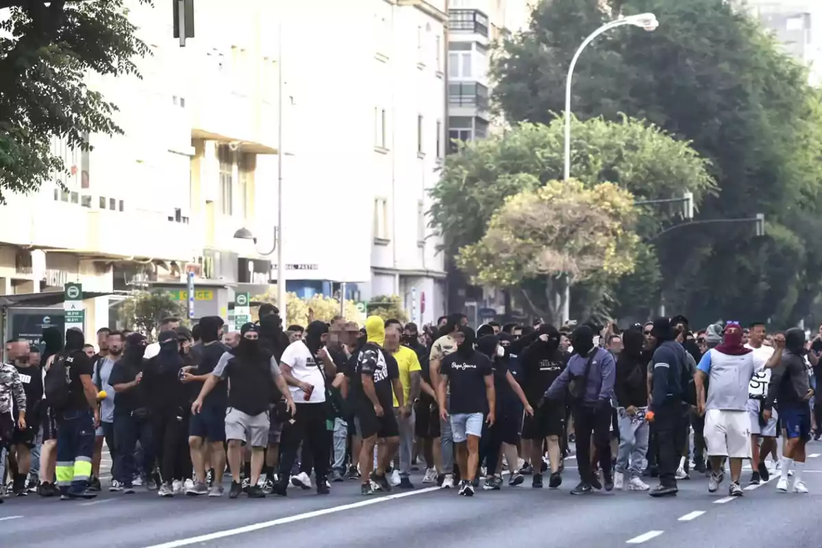 Un grupo numeroso de personas, muchas con el rostro cubierto, camina por una avenida en una ciudad rodeada de edificios y árboles. Un grupo numeroso de personas, muchas con el rostro cubierto, camina por una avenida en una ciudad rodeada de edificios y árboles.