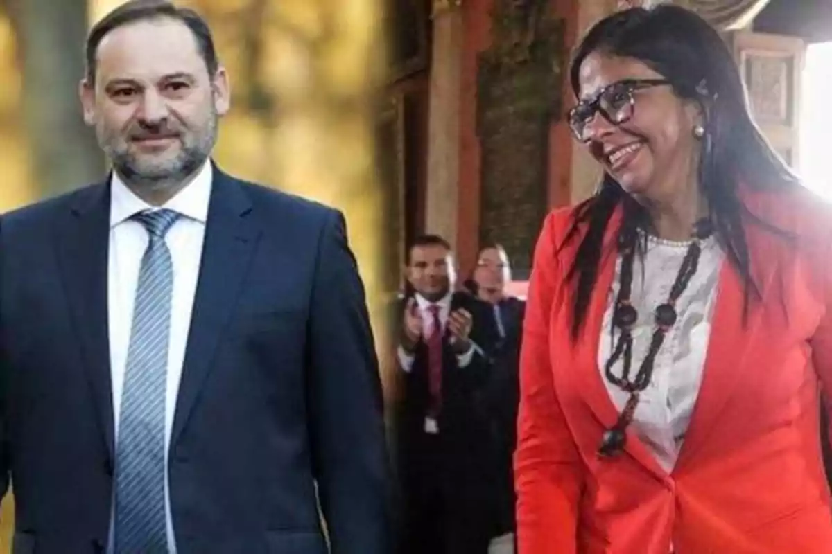 Dos personas vestidas formalmente, una con traje oscuro y corbata y otra con chaqueta roja y collar, en un entorno interior con personas aplaudiendo al fondo.