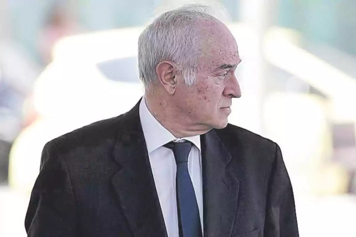 Un hombre mayor con cabello canoso y traje oscuro mira hacia un lado en un entorno exterior. Un hombre mayor con cabello canoso y traje oscuro mira hacia un lado en un entorno exterior.