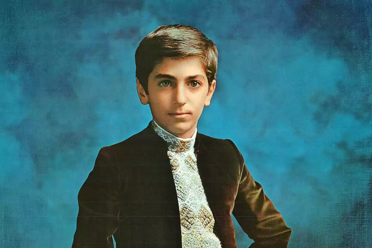 Retrato de un niño con cabello corto y oscuro, vestido con un traje elegante de terciopelo oscuro y camisa blanca decorada, posando frente a un fondo azul.