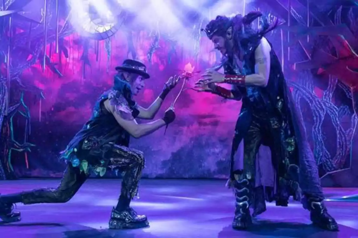 Dos actores en un escenario con iluminación púrpura y vestuario extravagante, uno de ellos arrodillado ofreciendo una flor al otro.