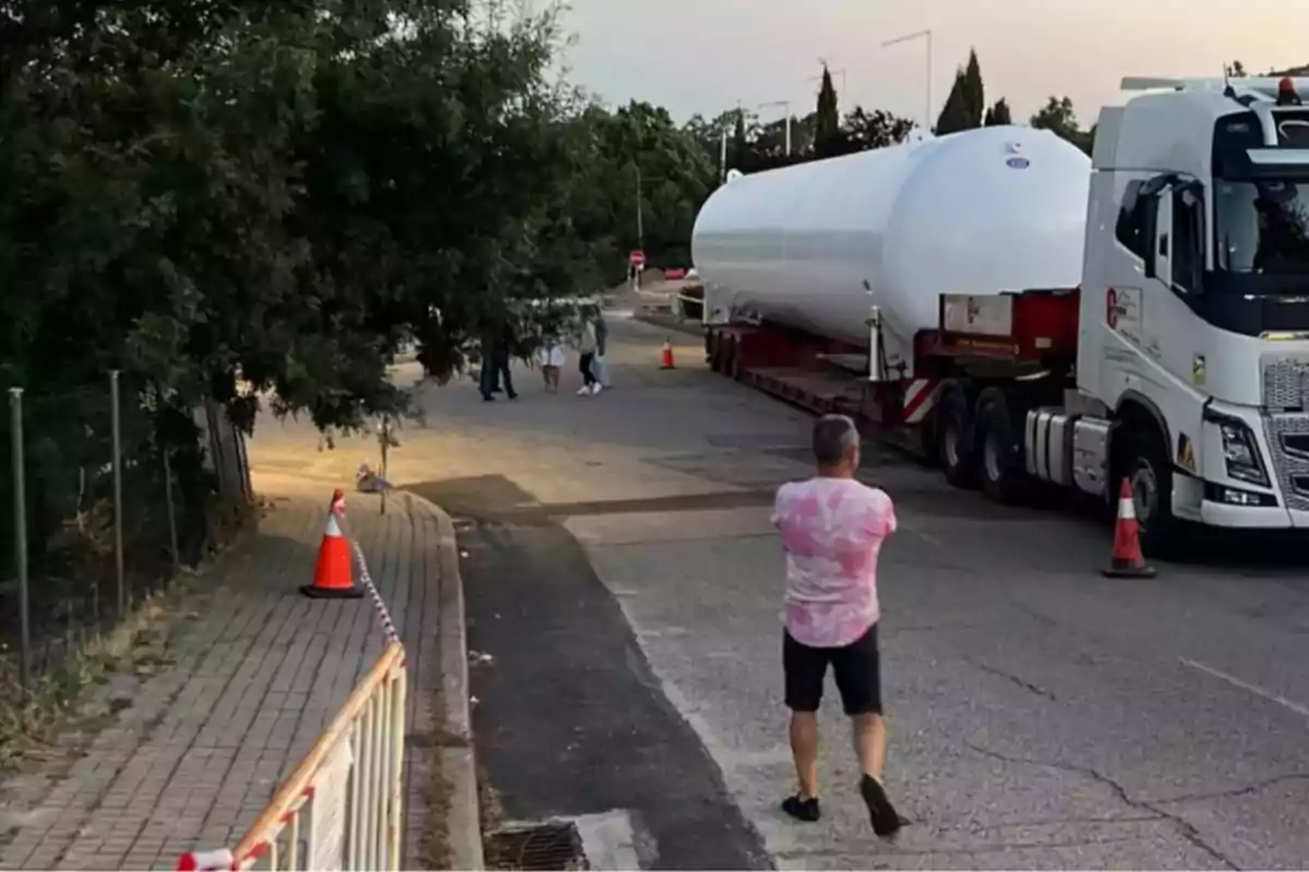 Un hombre observa un camión de gran tamaño que transporta un tanque blanco por una calle, mientras varias personas caminan al fondo y hay conos naranjas señalizando el área